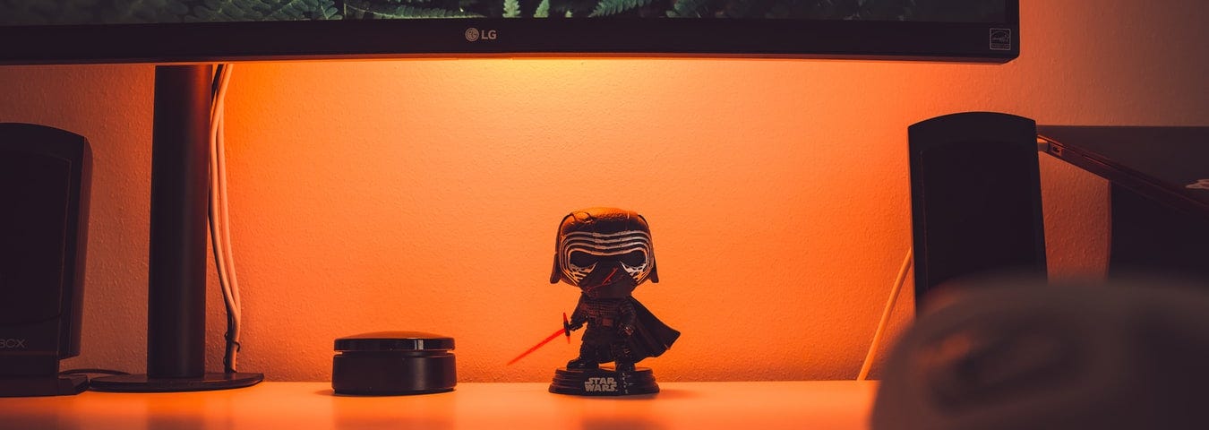 Uma estação de trabalho com um boneco do Kylo Ren embaixo do monitor, iluminado por uma luz laranja