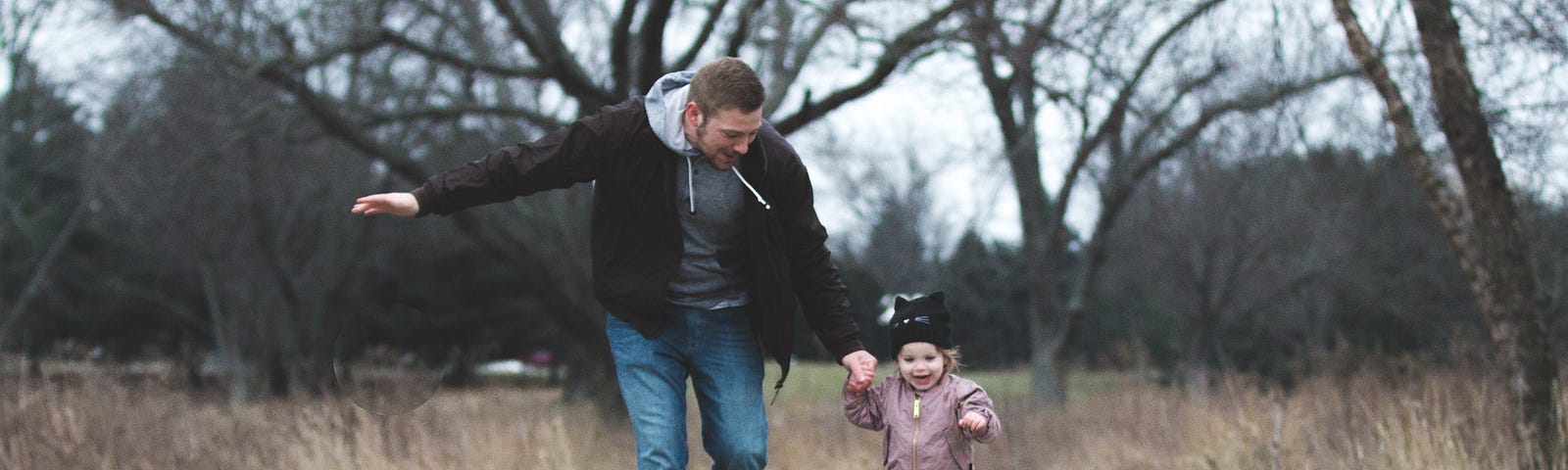Pai corre de mãos dadas com a filha bebê em um bosque repleto de árvores. A criança sorri, feliz. Esta foto ilustra um texto sobre como obter residência permanente na Estônia.