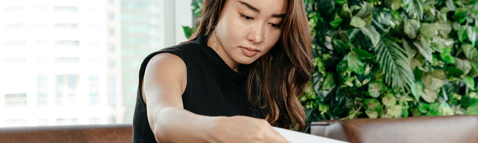 A woman wearing a black blouse and brown skirt is sitting on a brown leather couch in front of a window and a large plant with green foliage. She is looking through paperwork.