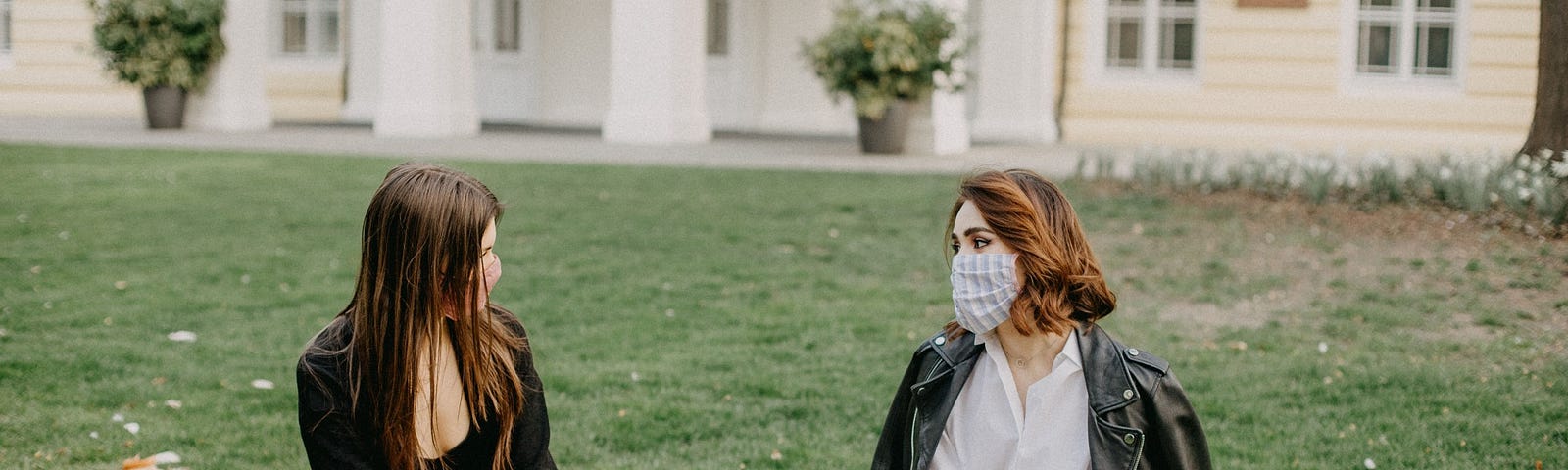 Two friends sit in a park and socialize with their masks on.