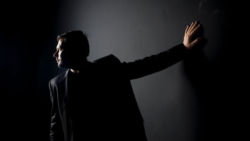 Image: A man in dark clothes and dim light, hand against the wall and looking the other way.