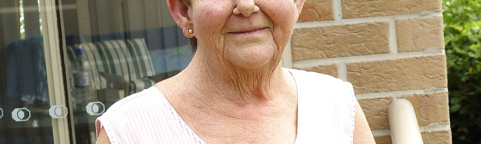 Elderly woman looking at the camera with a gentle smile on her face. One hand rests on a balcony railing and a plant with a red flower is in the forefront.