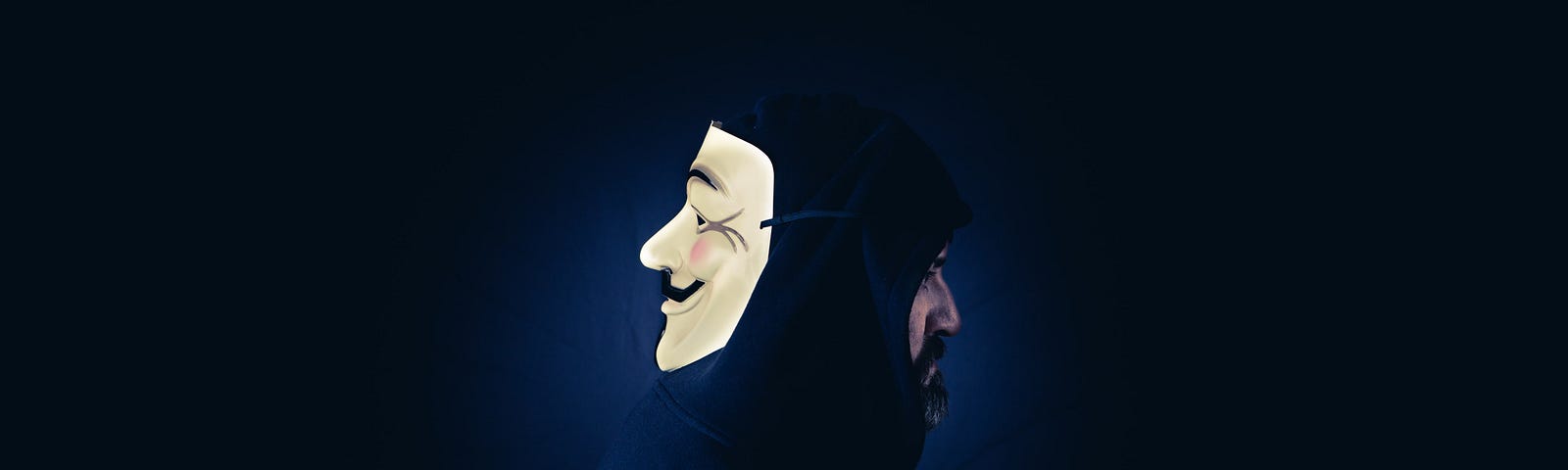 A man in a hoodie wears a Guy Fawkes mask on the back of head.