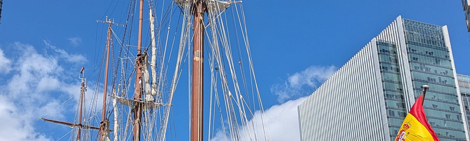 A docked 4-masted tall ship with Spanish flag at stern against a blue sky with white clouds