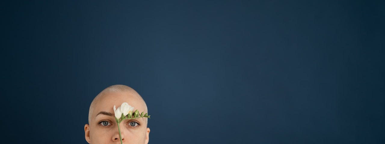 A bald woman with vibrant blue eyes holding a freesia flower to her forehead.