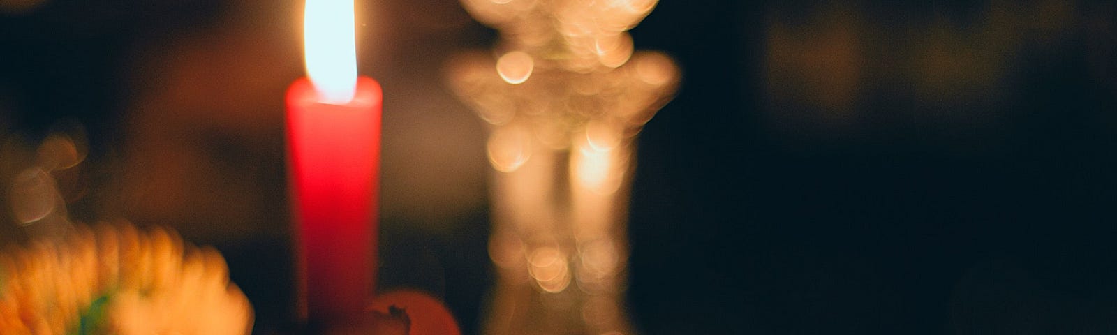 Photo of a golden chalice on a dark surface. Behind it, the image blurs, showing a lit, red tapered candle in candleholder, and an ornate crystal carafe with a stopper. A few other items are blurred and in the background, without being clearly identified. The darkness of the photo feels intimate, sacred, or of private importance.