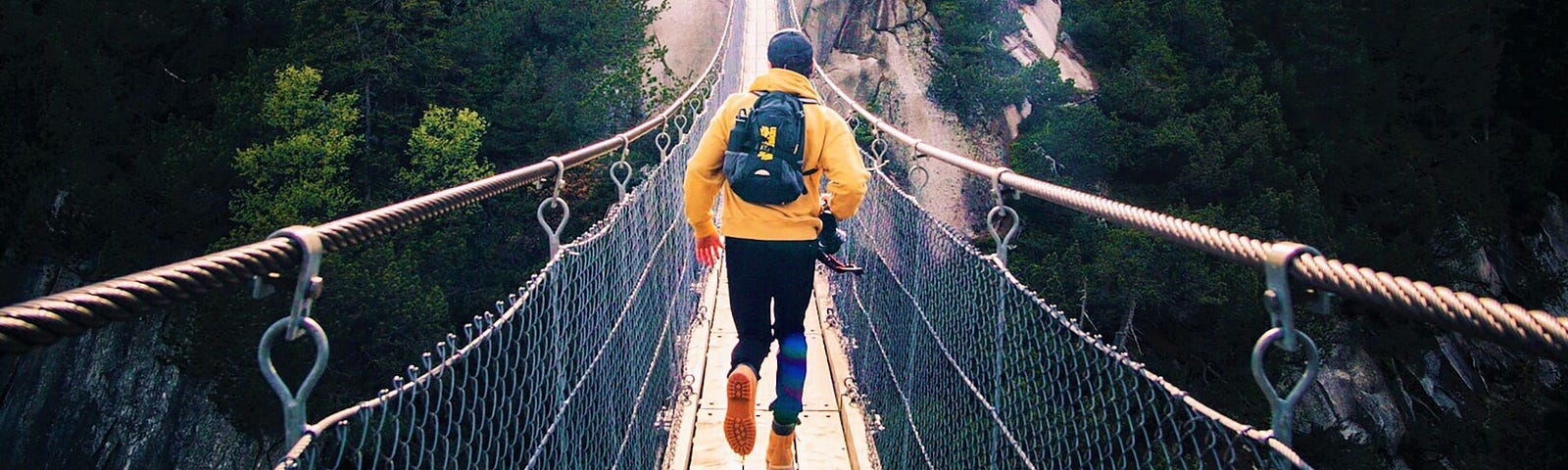back view of man wearing yellow jacket and backpack running across suspension bridge
