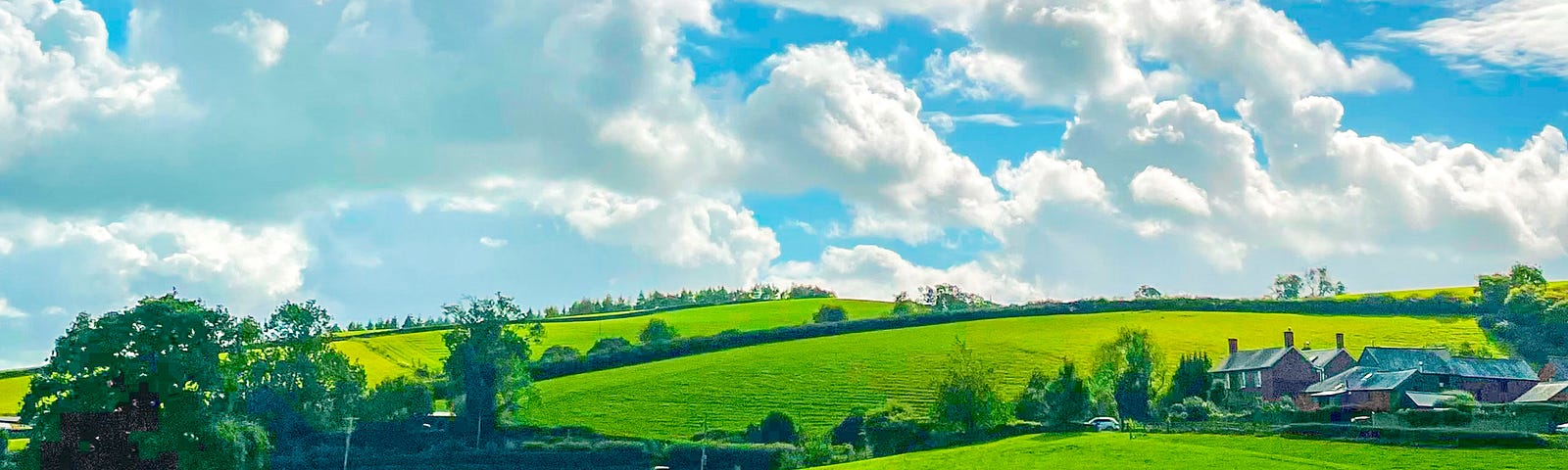 Green rolling hills with a red farmhouse in the distance, sheep in the forground, and the sky is full of white fluffy clouds.