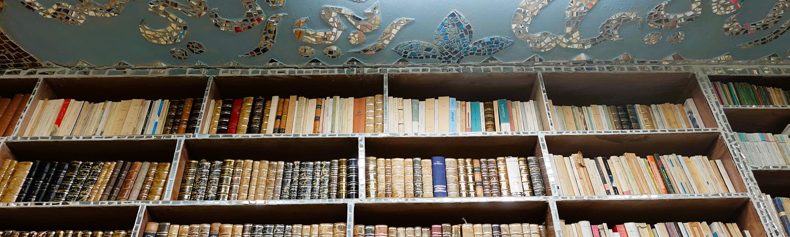 A curated bookshelf with classically bound tomes against a light blue ceiling decorated with mirror mosaics