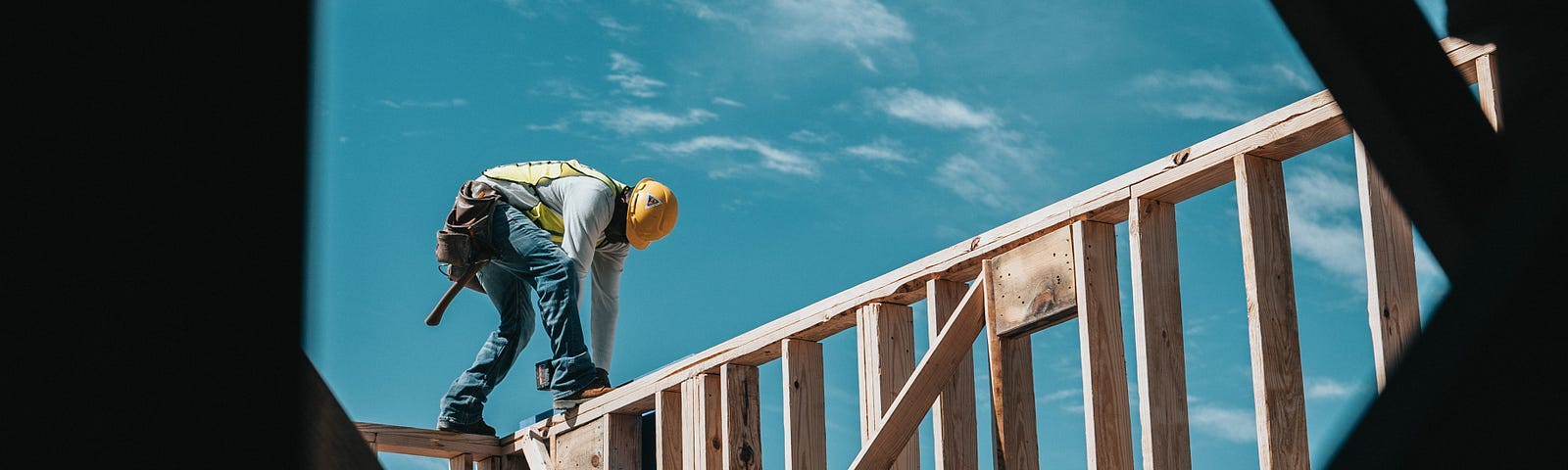 A construction worker wearing  jeans, a long sleeved light colored shirt, vest, hard hat, and tool belt stands atop a wooden frame of a building or house. They are bent over and working with a tool. Cross beams that are part of the construction are visible in the foreground of the picture.