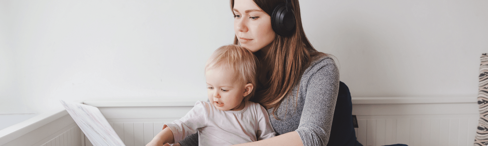 Woman sitting crossed legged on the floor, wearing a headset. She’s balancing a laptop on one knee and she has a baby sitting on her lap while she’s trying to type.