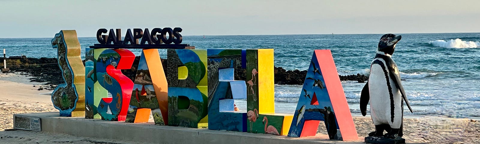 Tall, colorful letters on a white sandy beach spell ISABELA, with a statue of a Galápagos penguin statue on the side.