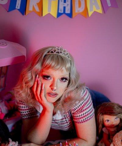 Young woman wearing a tiara and surrounded by dolls and a birthday cake.