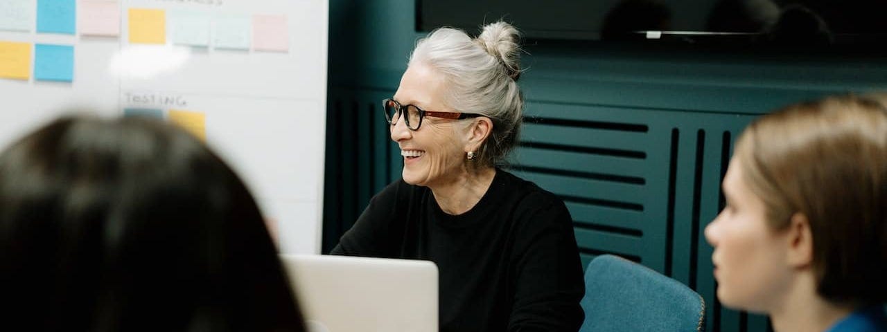 an older woman sits at a round table with a white board behind her, smiling in a direction off camera after transitioning from sex educator to sex coach.