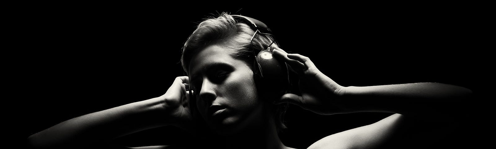 Black and white photo. Head and shoulders of a beautiful woman with headphones on emerges from a black background.