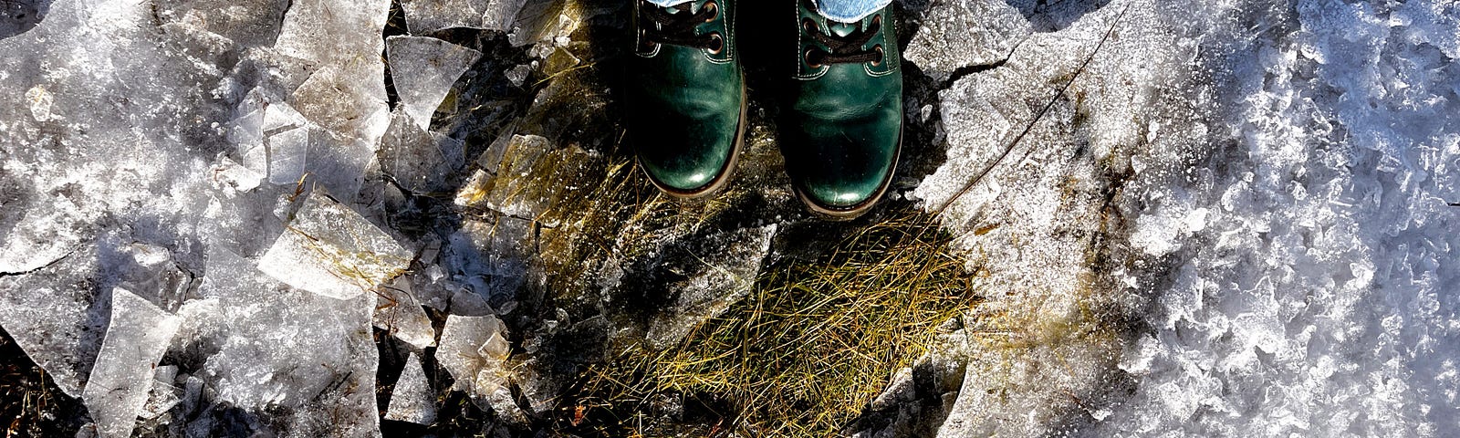 Selfie, looking down onto green lace-up boots, pale blue jeans and the bottom edge of a khaki-green coat. Standing on a small mound of mossy grass surrounded by the cracked remains of a layer of ice. Lit by cool winter sun.