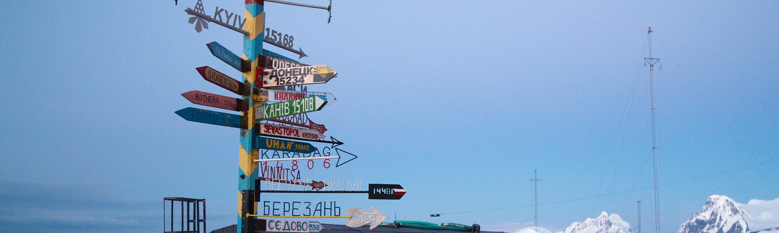 Black shack next to water, snow in the background and a blue and yellow pole in the foreground with signs pointing in different directions and S on top indicating South.