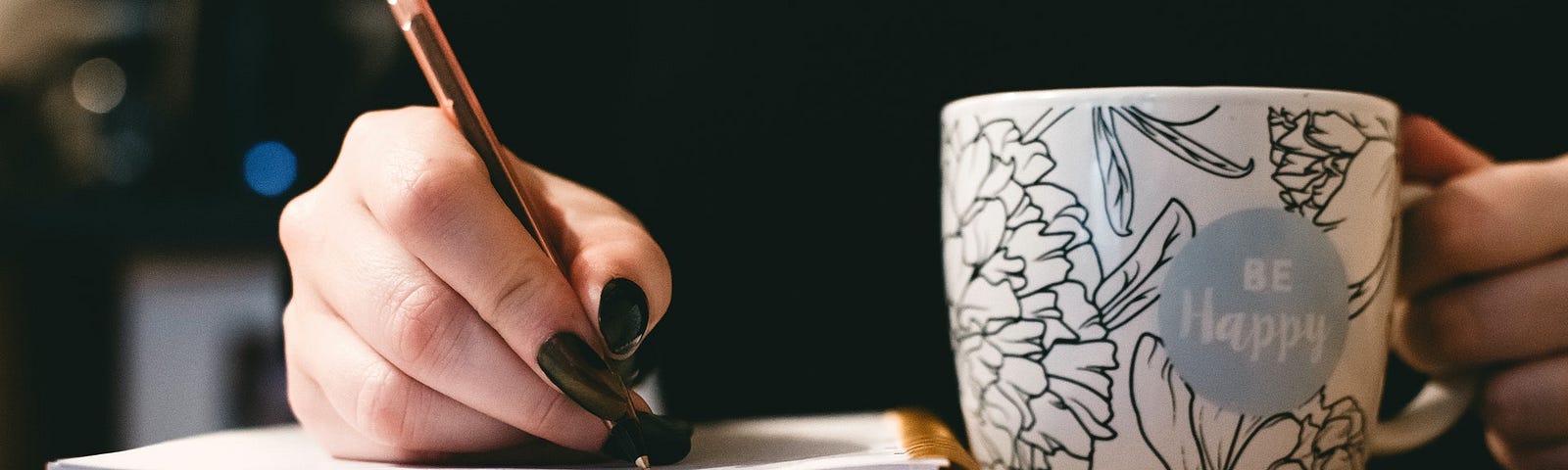 A person with long painted nails writes in their notebook. A floral mug with the words “Be Happy” rests in their other hand.