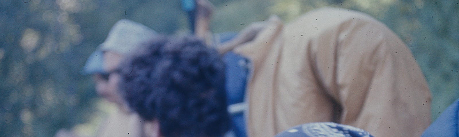 Teenage irl with long brown hair wearing a bandnana scarf and carrying a back pack with a coat over it, looking back at the camera with two other backpakers behind her.