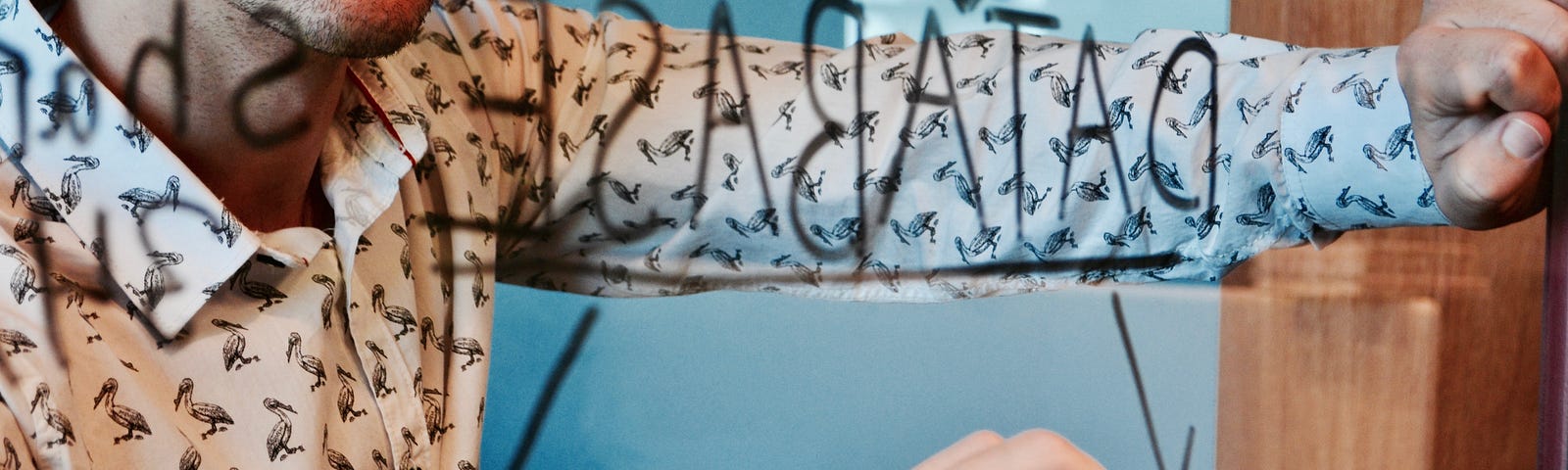 A man wearing a button-down bird shirt writes the word ‘database’ on a mirror with a black marker.