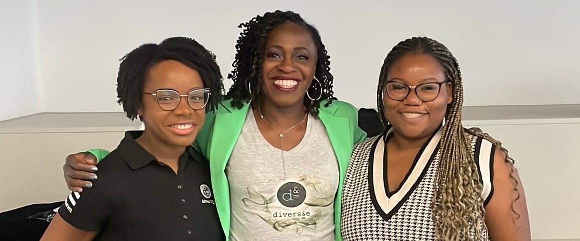 Three black women, with their arms around each other, stand side by side and are smiling to camera.