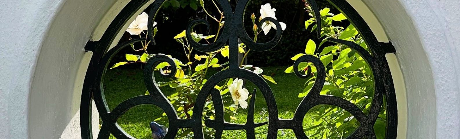 An ornate black wrought iron grille with the letter ‘H’ in the centre set into a round portal in a white fence. White rose bushes are in the garden behind the portal. The words, ‘Portal to Tranquility’, encircle the portal.