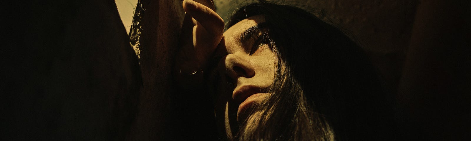 A girl with long brown hair is looking through a small gap in a prison cell that lets in some light.