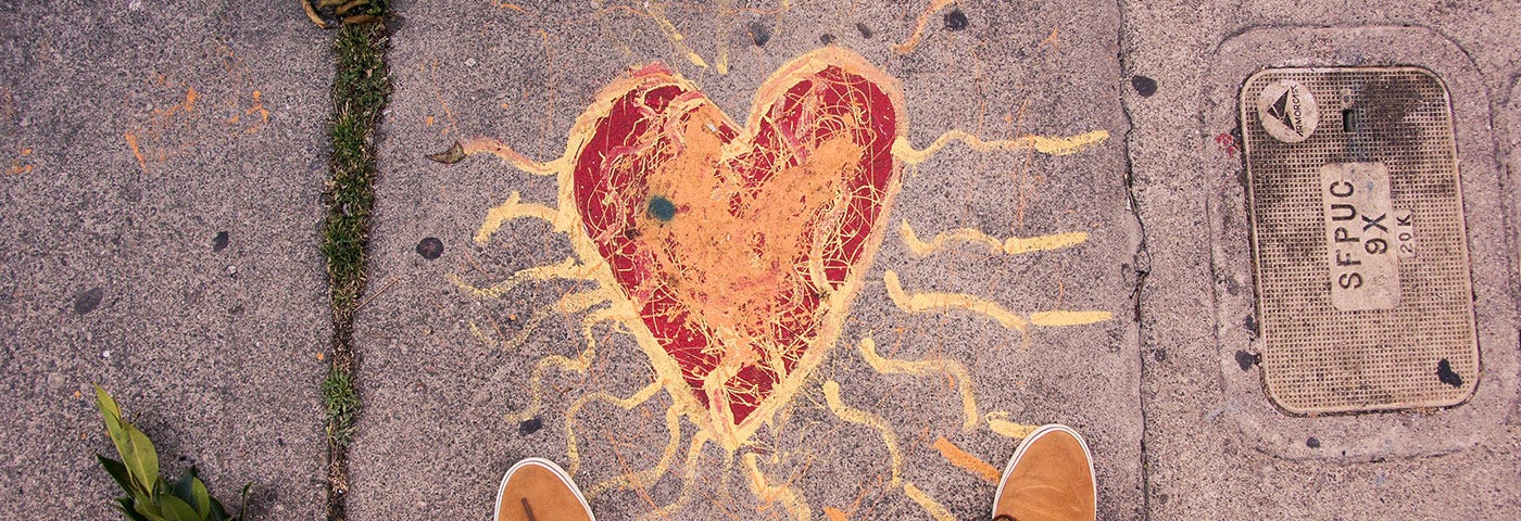 A red heart with yellow lines radiating from it draw in chalk on pavement with person’s shoes visible at the bottom of the frame.
