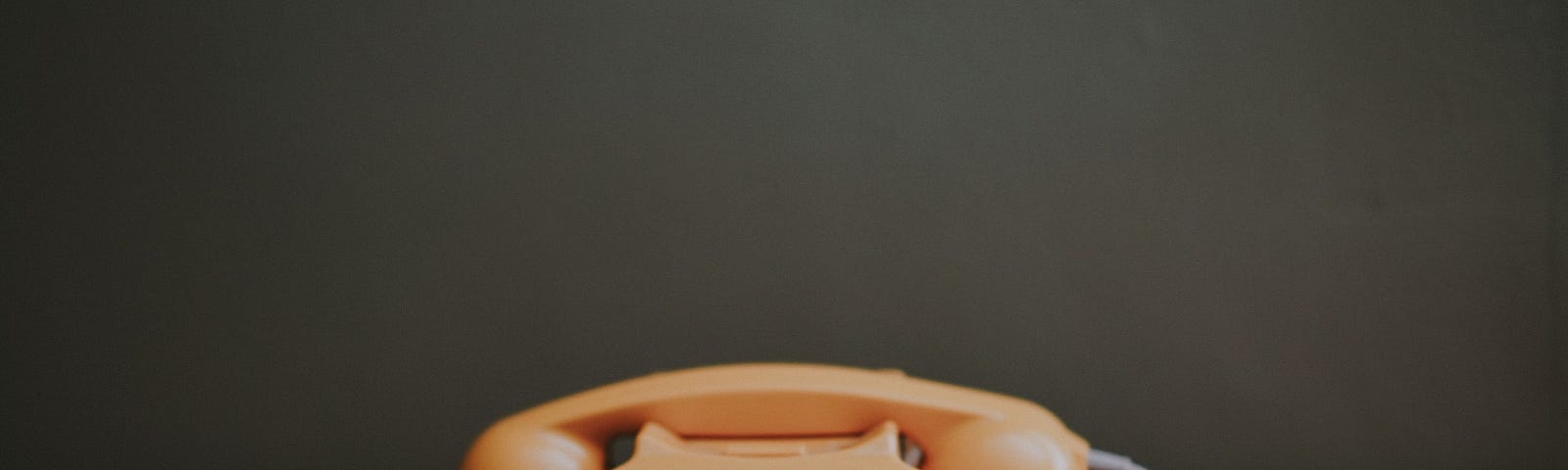 An orange rotary phone sitting on a wooden table in front of a dark gray-green wall