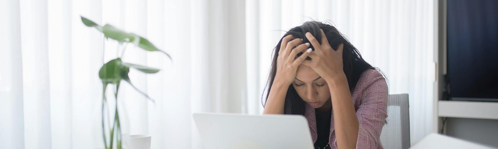 Image of woman in an office environment with her head in her hands in front of a laptop. She appears stressed.