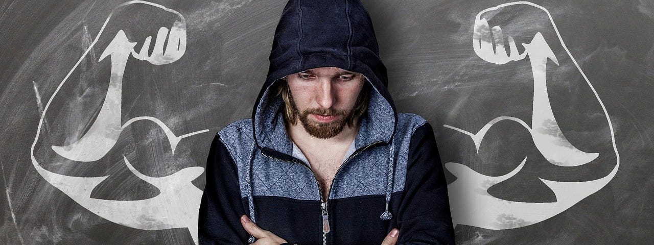 A man in severe emotional pain, leaning against a blackboard at his back, arms folding across his chest, gaze to the floor. Perfectly drawn on each side is a pair of arms, muscles bulging and flexed