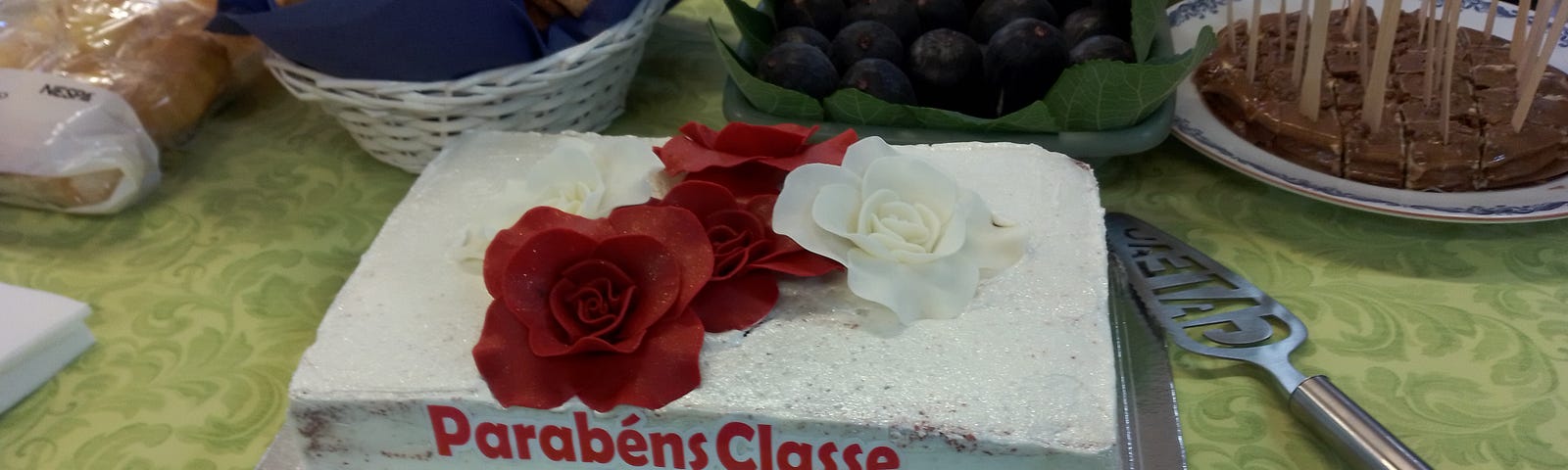 Buffet table with food. A white frosted cake with red roses that says “Parabens Classe 2023”