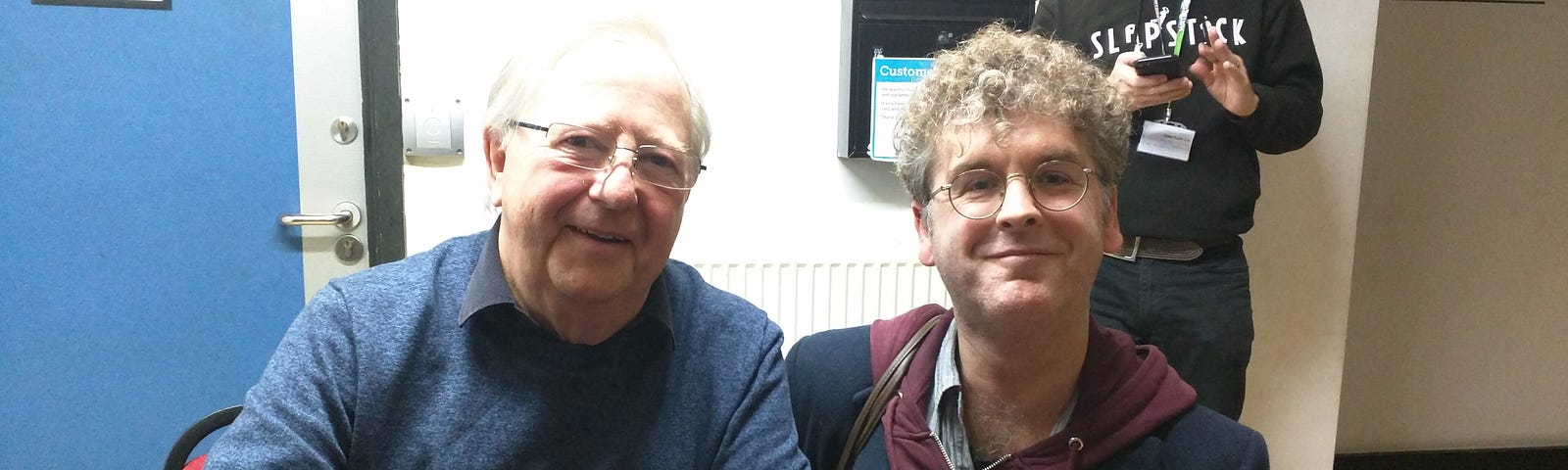 Comedian, Tim-Brooke-Taylor, in blue jumper and at signing table, poses with fan, Mike Hickman (author of this piece!) at the 2020 Slapstick Festival.