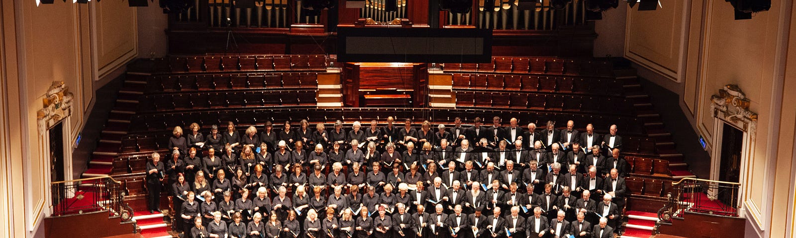 A symphony in a large concert hall with a choir standing behind. Photo presumbaly taken from a balcony in the audience seats.