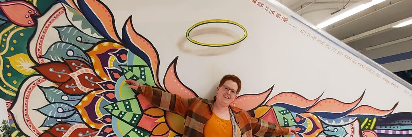 Photo of author, a redheaded woman wearing an orange shirt and brown snakeskin patterned pants, standing against a wall that is painted to look like she has huge variegated wings and a gold halo.