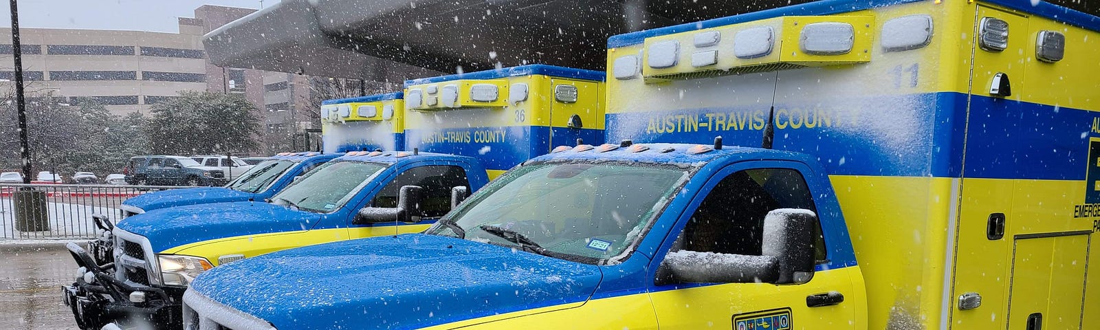 Two ambulances are parked outside a station during a snow storm.