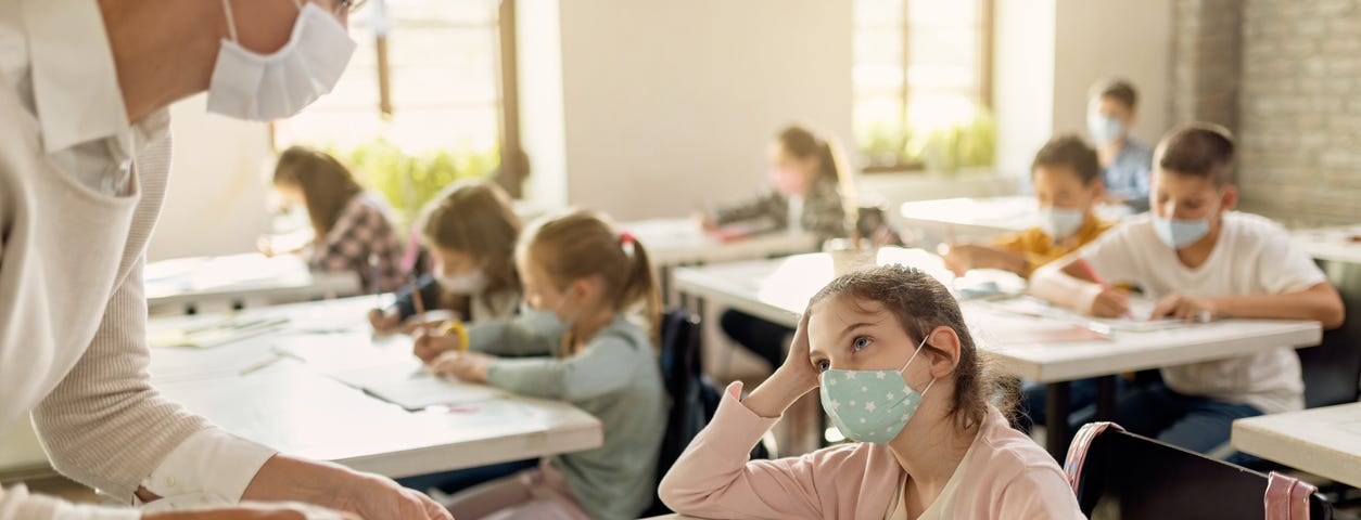 Small girl and her teacher wearing protective face masks while talking about exam papers curing a class at elementary school.