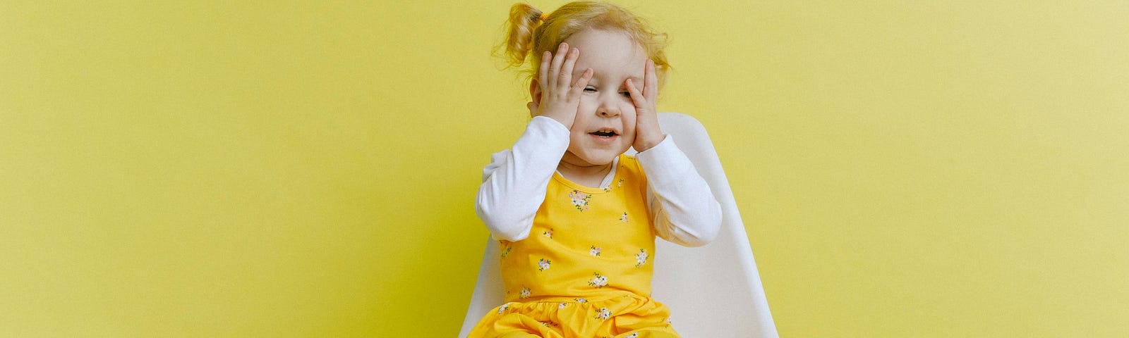 Little, blond girl sitting on a chair, yellow backdrop. Her hands on her face.