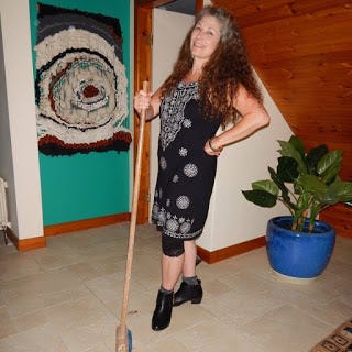 author in a black dress holding a broom with one hand, the other on her hip