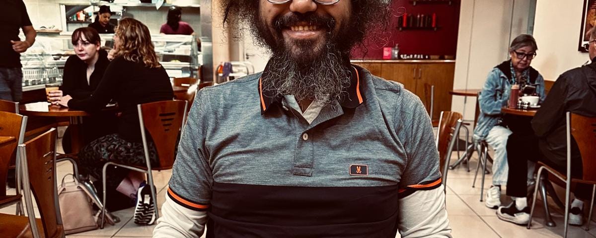 A man with curly hair and a beard, wearing glasses and a striped polo shirt, smiling at the camera. He is seated at a table with a full English breakfast in front of him.