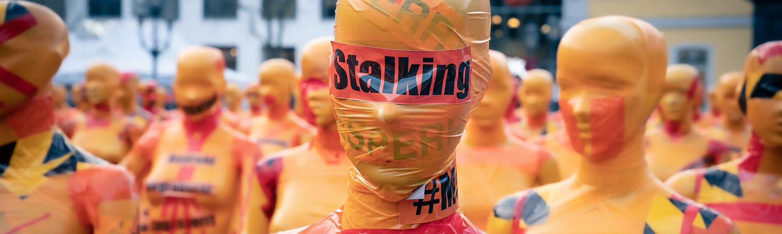 A series of mannequins wrapped in yellow tape, one with STALKING written on it. Josh Gets Busted by Jim Latham