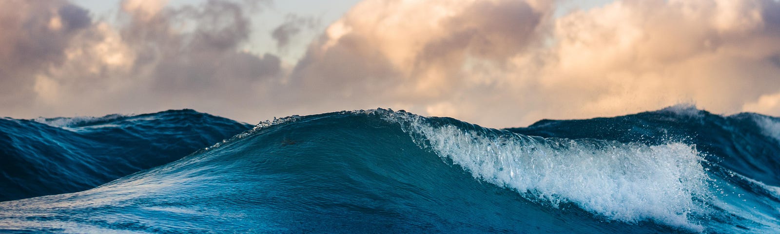 A beautiful crystal clear blue ocean wave rising, with a background of beautiful fluffy clouds in the distance.