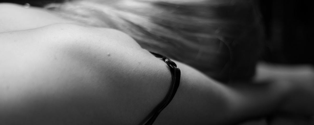 A black and white image of a woman lying on a bed. Only her head and shoulders are seen; she is implied to be naked. Her face is turned away from the camera.