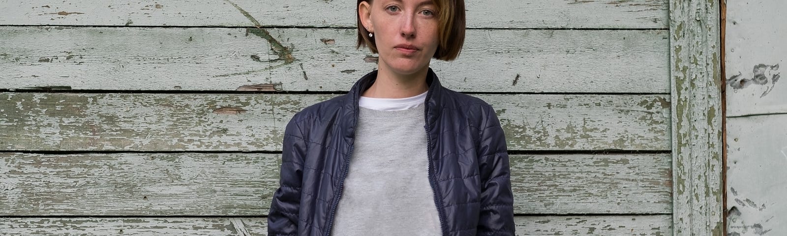 A woman with short hair stands against a faded wood-panel wall.