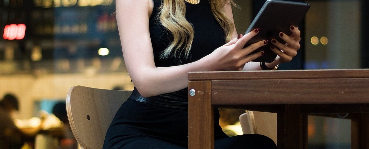woman, model dressed in black, long hair, sits at a table holding a tablet, electronic device, cafe background