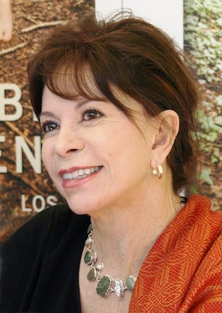 Isabel Allende is smiling and looking into the distance. She is wearing a black top, partially covered by an orange scarf.