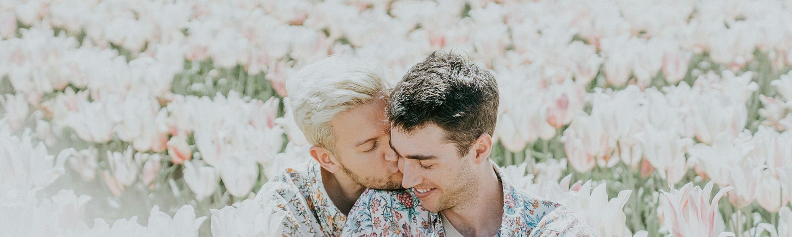 A man kisses the neck of another man as they sit in a field of flowers