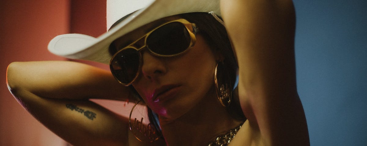 A woman photographed from mid-chest up, in a way which implies she is naked. Her arms are raised, apparently fastening or unfastening a heavy gold chain around her neck. She is wearing a cowboy hat, gold-rimmed sunglasses, and large, gold, hoop earrings.