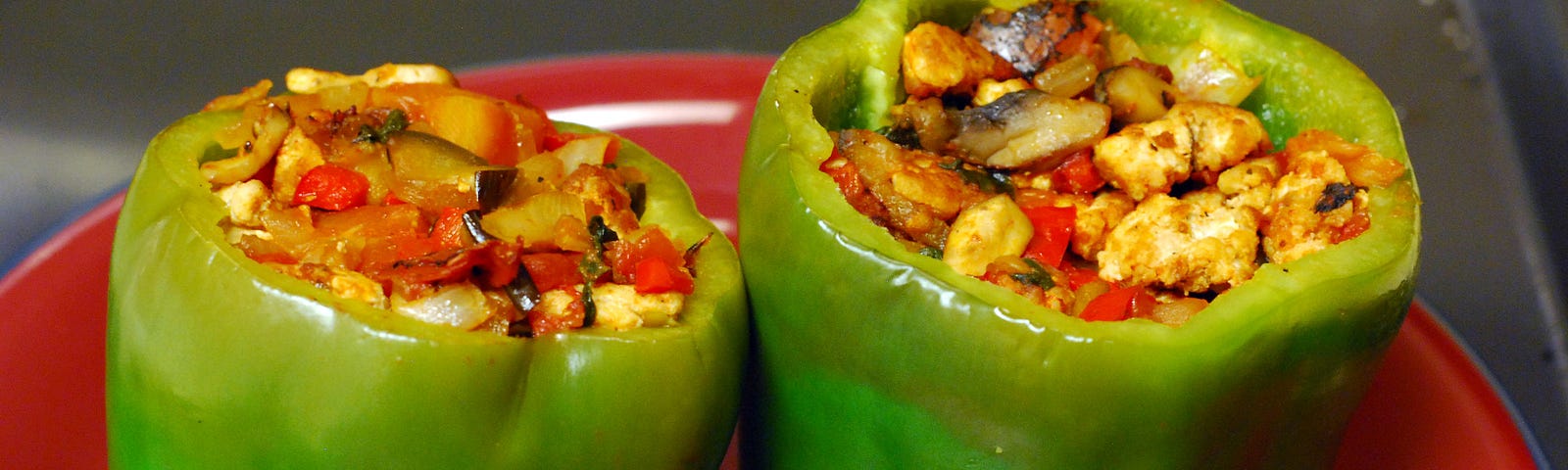 Two uncooked green capsicums (bell peppers) filled with a mixture of chopped vegetables, standing on a red plate.
