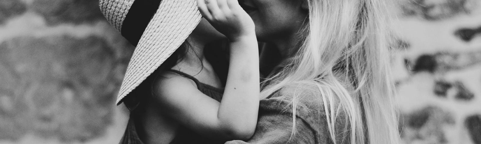 Black and white photo of mom holding daughter close to her face with little girl holding onto her hat and wrapping it in towards her mom.